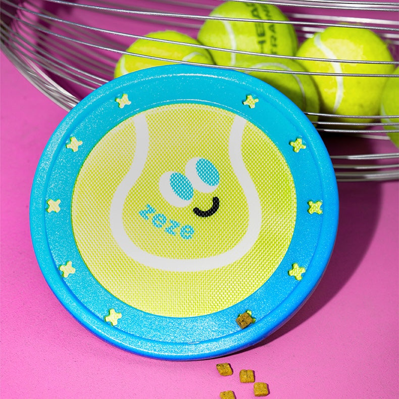 Round yellow and blue toy with a tennis ball design and '2020' text, placed on a pink surface with tennis balls in the background.