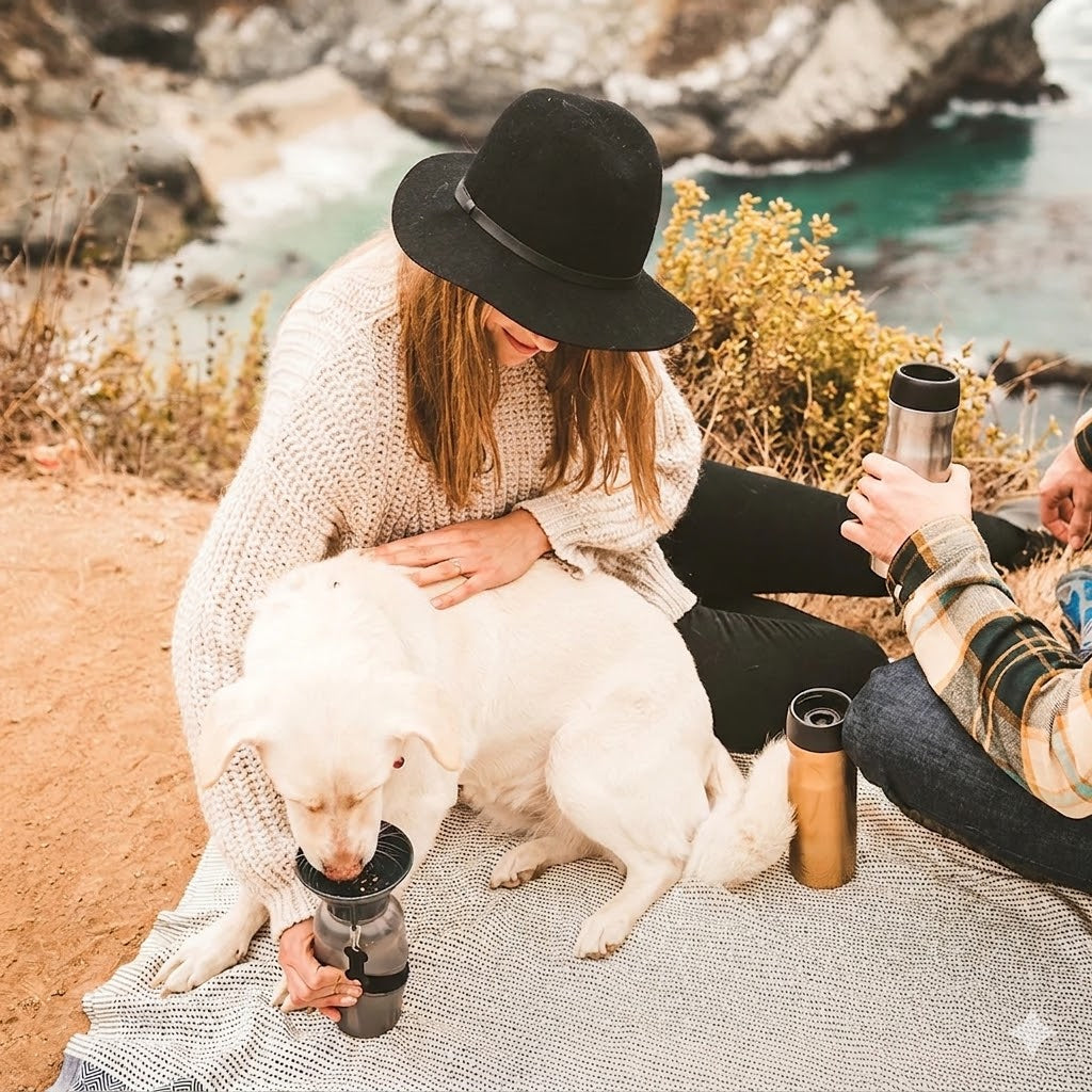Woman with a black hat and white dog sitting by a rocky coastline, holding a thermos.